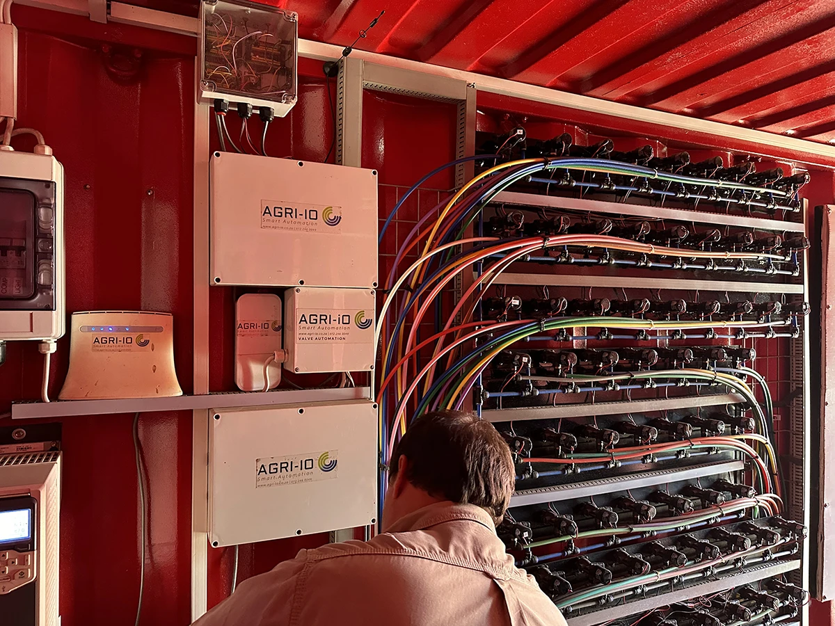 The image shows a man working with Agri.io inside a control unit of an automated farming system. The interior is filled with a series of electrical components, wiring, and devices labeled AGRI-IO which appear to be part of a smart automation system for agricultural management.