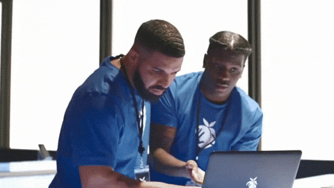 Two individuals wearing blue shirts collaborate while working on a laptop. One person types on the keyboard while the other looks over his shoulder, both focused on the screen.